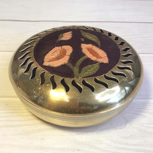 Peut inclure: Boîte à bijoux ronde en laiton avec un motif floral sur le couvercle. Le couvercle présente un fond violet foncé avec deux fleurs couleur pêche et des feuilles vertes. La boîte a des découpes décoratives sur le bord.