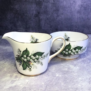 Peut inclure: Service à crème et sucrier en porcelaine blanche. Le pot à crème a une anse et un bec verseur, et le sucrier a un pied. Les deux pièces sont décorées d'un motif floral avec des fleurs blanches, des feuilles vertes et une bordure dorée.