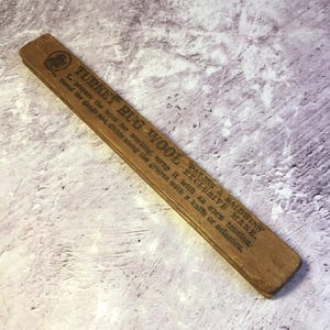 Könnte beinhalten: Ein Vintage-Holz-Türkei-Teppichwollmaß mit aufgedrucktem Text. Das Maß ist rechteckig und braun, mit dem Text "TURKEY RUG WOOL" und Anweisungen. Eine Truthahn-Grafik ist sichtbar. Das Maß wird zur Vorbereitung von Wolle verwendet.