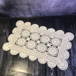 Puede incluir: Camino de mesa rectangular de ganchillo color crema con borde festoneado. El centro presenta un intrincado patrón calado de motivos florales. El camino de mesa está sobre una superficie de madera oscura.