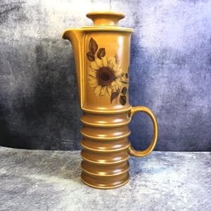 Puede incluir: Jarra de cerámica de color amarillo mostaza con un diseño de girasol. La jarra tiene un cuerpo alto y acanalado, un asa curva y una tapa. El girasol está representado con pétalos marrones y amarillos y hojas verdes. Una pieza decorativa para la cocina.