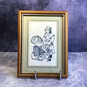 Puede incluir: Una impresión en blanco y negro enmarcada de una niña con una muñeca en un cochecito vintage. La impresión está en un marco de madera de color dorado y se exhibe en un soporte plateado.