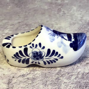 Może przedstawiać: Biały ceramiczny holenderski chodak z niebieskimi wzorami kwiatowymi i geometrycznymi. Ozdobne obuwie ma zaokrąglony nosek i puste wnętrze. Niebieskie wzory obejmują stylizowane kwiaty i motywy liści, typowe dla ceramiki z Delft.