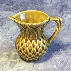 Peut inclure: Un petit pot à crème en céramique brun avec un motif de chardon. Le pot à crème a une poignée et un corps arrondi.