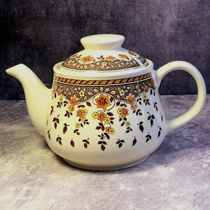 Könnte beinhalten: Cremefarbene Keramik-Teekanne mit Deckel, Ausguss und Henkel. Die Teekanne ist mit einem Blumenmuster in Orange-, Gelb- und Brauntönen verziert. Das Design umfasst Blumen und Blätter. Die Teekanne ist im Vintage-Stil.