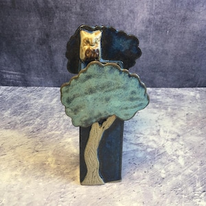 Könnte beinhalten: Keramikskulptur eines Baumes mit einer Eule. Der Baum hat eine blau-grüne Krone und einen beigefarbenen Stamm. Die Eule ist beige und weiß. Die Skulptur steht vor einem dunkelblauen Hintergrund.