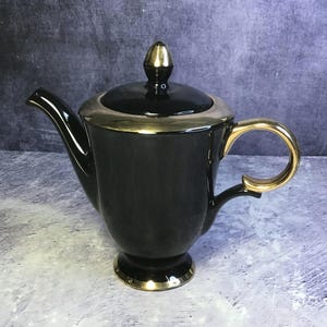 Könnte beinhalten: Schwarze Keramik-Teekanne mit goldenen Verzierungen am Rand, Griff und Boden. Der Deckel hat einen goldenen Knauf. Der Ausguss ist gebogen und der Griff ist eine große, offene Schlaufe. Die Teekanne steht auf einem kleinen, runden Sockel.