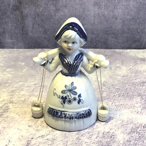 Könnte beinhalten: Keramikfigur eines holländischen Mädchens in traditioneller Kleidung. Das weiße Kleid ist mit blauen Blumenmustern und Verzierungen versehen. Sie trägt zwei kleine Eimer, die an Schnüren von ihren Armen hängen. Hut und Oberteil der Figur sind blau akzentuiert.