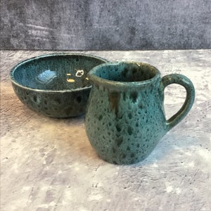 Puede incluir: Un juego de cuenco y jarra de crema de cerámica verde moteada. El cuenco y la jarra de crema son de un verde azulado oscuro con un esmalte moteado.