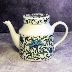 Könnte beinhalten: Weiße Keramik-Teekanne mit Deckel, verziert mit einem floralen Muster in Blau- und Grüntönen. Die Teekanne hat einen gebogenen Ausguss und einen Henkel. Das florale Design bedeckt den Körper und den Deckel.