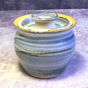 Puede incluir: Una mantequera de cerámica hecha a mano con tapa. El plato es cilíndrico con un patrón de rayas azul claro y marrón. El borde del plato y la tapa son amarillos. La tapa tiene un pequeño asa elevada.