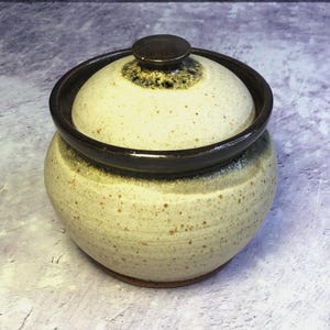 Puede incluir: Un tarro de cerámica hecho a mano con tapa. El tarro es de color beige moteado con detalles marrones alrededor del borde y la tapa. La tapa tiene un pequeño pomo redondo. El tarro probablemente se utiliza para el almacenamiento.