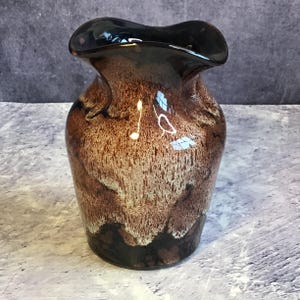 May include: A brown and white glazed ceramic vase with a wavy rim. The vase has a mottled brown and white glaze with a black rim.