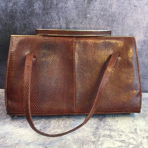 Puede incluir: Un bolso de mano de cuero marrón con un patrón de piel de serpiente y un cierre superior de metal plateado. El bolso tiene un solo asa superior y una correa de hombro desmontable.