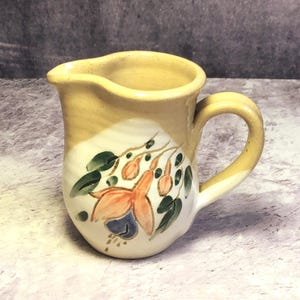 Puede incluir: Jarra de cerámica pequeña pintada a mano, con cuerpo crema y borde y asa amarillos. Decorada con un diseño floral con flores naranjas y azules y hojas verdes. La jarra tiene un asa curva y un pico.