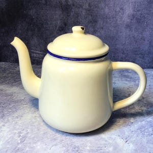 Könnte beinhalten: Eine cremefarbene Emaille-Teekanne mit rundem Körper, geschwungenem Griff und einem Ausguss mit abgeplatzter Spitze. Der Deckel hat einen kleinen Knauf und einen dunkelblauen Rand, der zum Akzent am unteren Rand des Deckels passt.