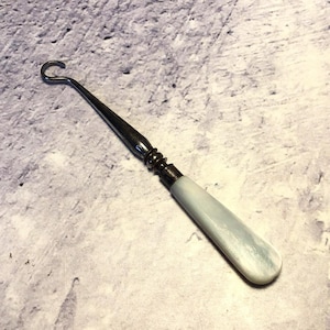 May include: A button hook featuring a polished silver-toned metal shaft with a curved hook at one end and a smooth, pale blue, marbled-effect handle at the other. The handle is cylindrical with a rounded end, connected to the metal by a ribbed ferrule.