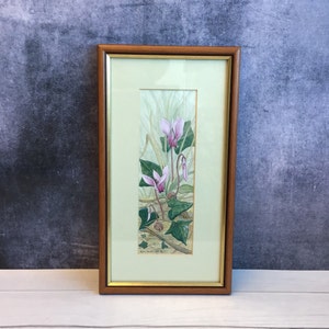 Puede incluir: Una pintura de acuarela enmarcada de flores de ciclamen rosas con hojas verdes. La pintura está sobre un fondo verde claro y está enmarcada en un marco de madera marrón claro.