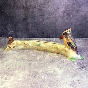 Puede incluir: Un jardinera de cerámica con forma de tronco, con un pájaro colorido. La jardinera es principalmente beige con detalles marrones, y el pájaro tiene un plumaje vibrante azul, amarillo y naranja. Hay detalles verdes en la base.