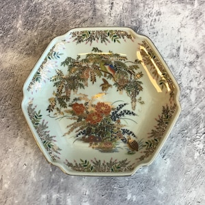 Può includere: Un piatto ottagonale in ceramica bianca con un bordo dorato e intricati disegni floreali e di uccelli. Il piatto presenta un delicato motivo floreale con accenti dorati e due uccelli appollaiati su un ramo.
