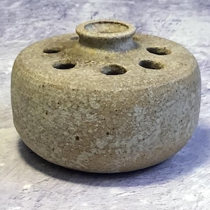Puede incluir: Un tope de flores redondo de cerámica beige con una abertura central y seis agujeros circundantes. La superficie texturizada tiene un aspecto moteado, y el tope está diseñado para sujetar los tallos de las flores en arreglos florales.