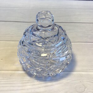 May include: A clear glass sugar bowl with a decorative, faceted design. The bowl has a round, domed lid with a knob on top.