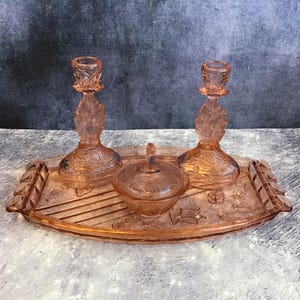 Könnte beinhalten: Ein Vintage-Schminkset aus rosa Glas mit einem Tablett, zwei Kerzenhaltern und einem kleinen Glas. Das Set hat ein zartes Blumenmuster.