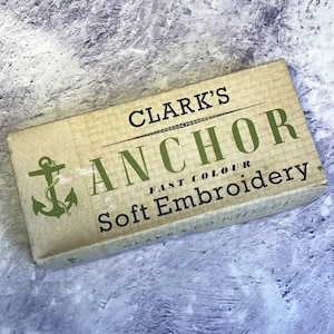 Könnte beinhalten: Eine rechteckige Schachtel mit Clark's Anchor Soft Embroidery Garn. Die Schachtel ist beige mit grünem Text und einem Ankerlogo. Der Text lautet "Clark's Anchor Fast Colour Soft Embroidery".