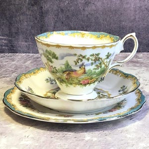 Puede incluir: Juego de taza y platillo de té vintage. La taza y el platillo de porcelana blanca presentan una escena de paisaje con un pájaro, árboles y flores. Los bordes están adornados con detalles dorados y turquesas, creando un aspecto elegante.