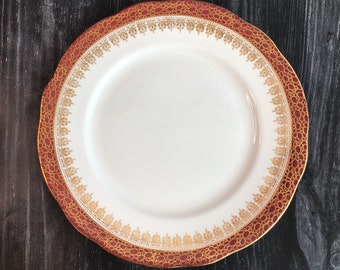 May include: A white ceramic plate with a red and gold rim. The plate has a decorative pattern of gold swirls and dots.