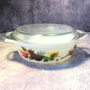 Op de afbeelding: Een witte ovenschaal van melkglas met een doorzichtig glazen deksel en geïntegreerde handgrepen aan de zijkanten. De buitenkant is voorzien van een kleurrijk groentepatroon met een paarse aubergine, rode radijs, groene erwten en diverse kruiden.
