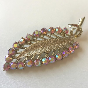 Op de afbeelding: Goudkleurige broche in de vorm van een blad met iriserende, ronde edelstenen. De broche heeft een gedetailleerd, getextureerd bladerontwerp met een steel en een decoratieve krul aan de bovenkant. De edelstenen zijn langs de randen van het blad geplaatst.