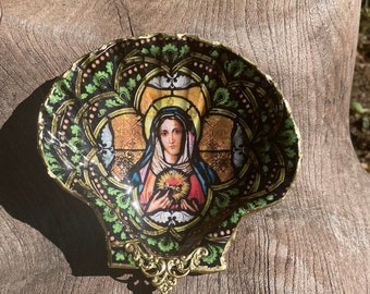 Puede incluir: Una concha decorativa con un diseño de vitral que representa a una mujer sosteniendo un corazón. La concha es dorada y verde con detalles intrincados.