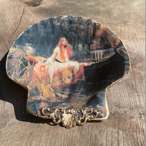 May include: A decorative seashell dish with a gold trim and a painted image of a woman in a boat. The woman is wearing a white dress and has long red hair.
