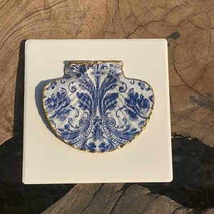 Op de afbeelding: Een witte keramische schelp met een blauw en gouden bloemenpatroon. De schelp is gemonteerd op een witte vierkante tegel.