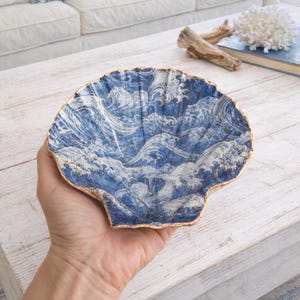May include: A decorative shell-shaped dish with a blue and white wave pattern and a gold rim. The dish is held in a hand and is on a white wooden table. The dish is a decorative item.