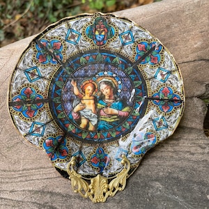 May include: A round, decorative plate with a stained glass design featuring a depiction of the Virgin Mary and baby Jesus. The plate has a gold-colored metal frame with ornate details.