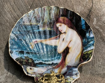 May include: A seashell with a painting of a mermaid inside. The seashell is decorated with a gold octopus charm.