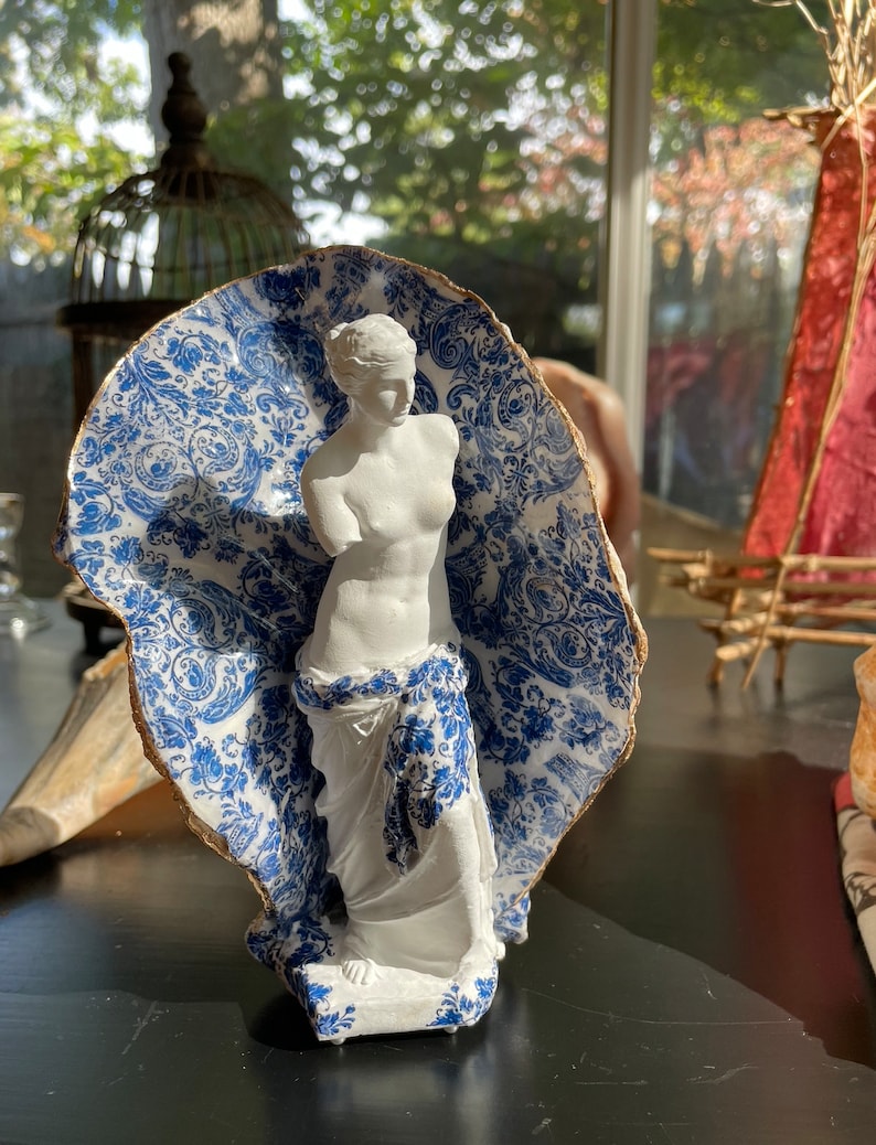Pu&ograve; includere: Una statua di donna in ceramica bianca, in piedi con un braccio alzato, &egrave; parzialmente coperta da un guscio di ceramica con un motivo blu e bianco. Il guscio ha un bordo dorato.