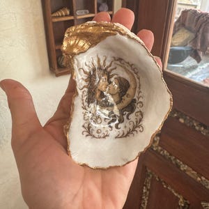 以下が含まれることがあります： 金色の縁と内側に絵が描かれた装飾的なカキの貝殻。内側には、太陽と月のモチーフを持つ、抱き合う2人の人物の詳細なイラストが描かれています。貝殻は手で持たれています。