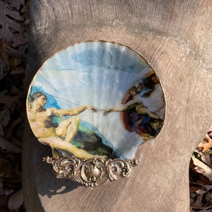 May include: A decorative seashell dish with a gold trim. The dish features a reproduction of Michelangelo's The Creation of Adam painting.