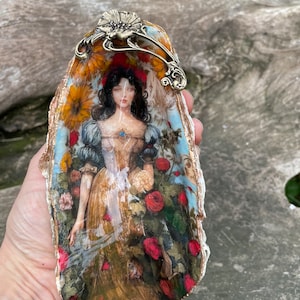 May include: A decorative seashell dish featuring a painted image of a woman in a garden setting. The woman is wearing a white dress with a blue sash and a red headband. The dish has a gold metal handle.