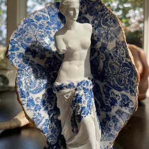 Pu&ograve; includere: Una statua in gesso bianco della Venere di Milo, in piedi in un guscio di porcellana blu e bianco con un bordo dorato.