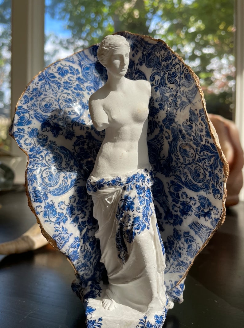 Pu&ograve; includere: Una statua in gesso bianco della Venere di Milo, in piedi in un guscio di porcellana blu e bianco con un bordo dorato.