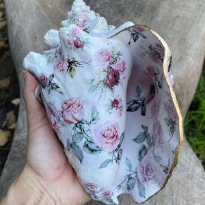 May include: A large white seashell with a gold trim, decorated with a floral pattern of pink roses and green leaves.