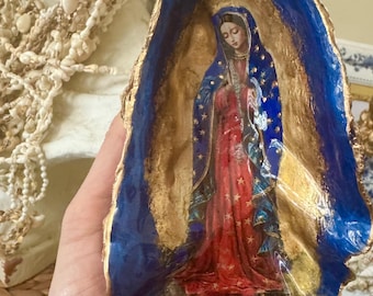 Puede incluir: Una obra de arte en concha de ostra que representa a la Virgen María. La obra de arte tiene un fondo dorado y un borde azul. La Virgen María lleva un vestido rojo y un velo azul con estrellas doradas.