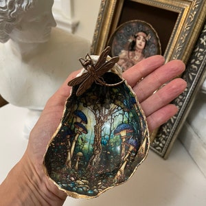 Puede incluir: Una concha de mar con un borde dorado, pintada con una escena de bosque con setas y árboles. Una libélula dorada está unida a la parte superior de la concha.