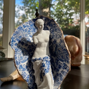 Pu&ograve; includere: Una statua in porcellana bianca di una donna in piedi in un guscio di porcellana blu e bianco. Il guscio ha un motivo floreale.