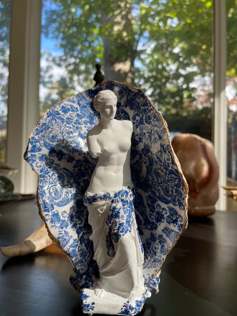 Pu&ograve; includere: Una statua in porcellana bianca di una donna in piedi in un guscio di porcellana blu e bianco. Il guscio ha un motivo floreale.