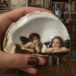 May include: A decorative oyster shell featuring a painted image of two cherubs. The cherubs have brown hair and small wings. The shell is white with a textured, brown edge. The artwork is reminiscent of classical paintings.